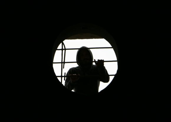 Silhouette of a person in a hoodie behind a round, barred window creating a mysterious scene.