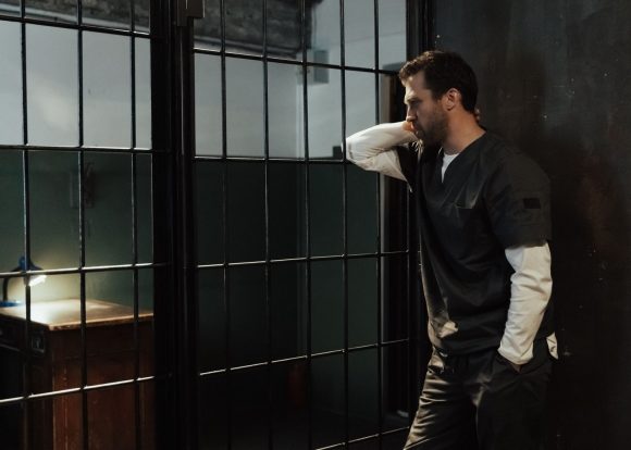 Man in uniform stands pensively by barred prison wall, illuminated by dim light.